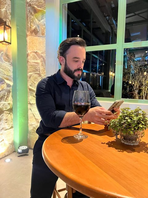 Felipe Ramalho em um jantar, olhando o celular à mesa, em ambiente iluminado à noite.